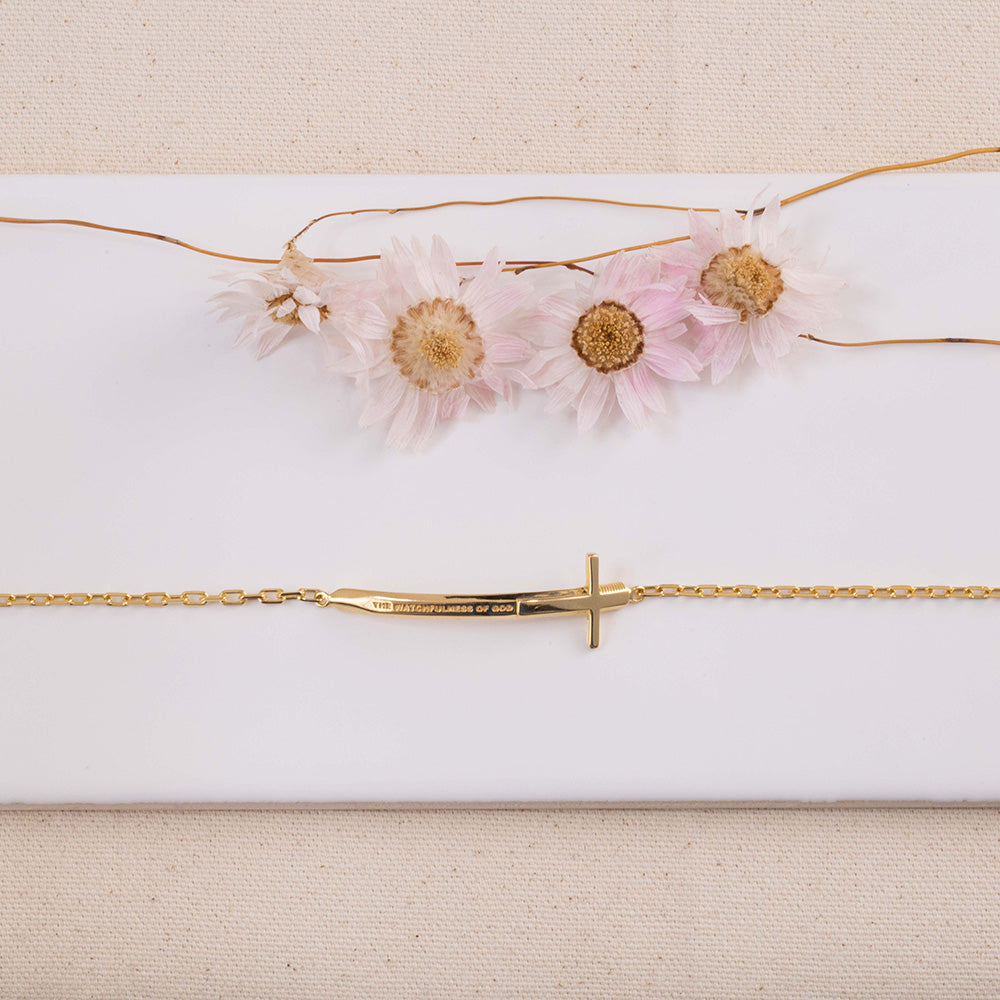 Pulseira Cristã Espada de Dois Gumes Feminino em Prata de Lei com banho de ouro 18kt em cima de azulejo branco com um fundo rosa bem claro e flores secas ao lado